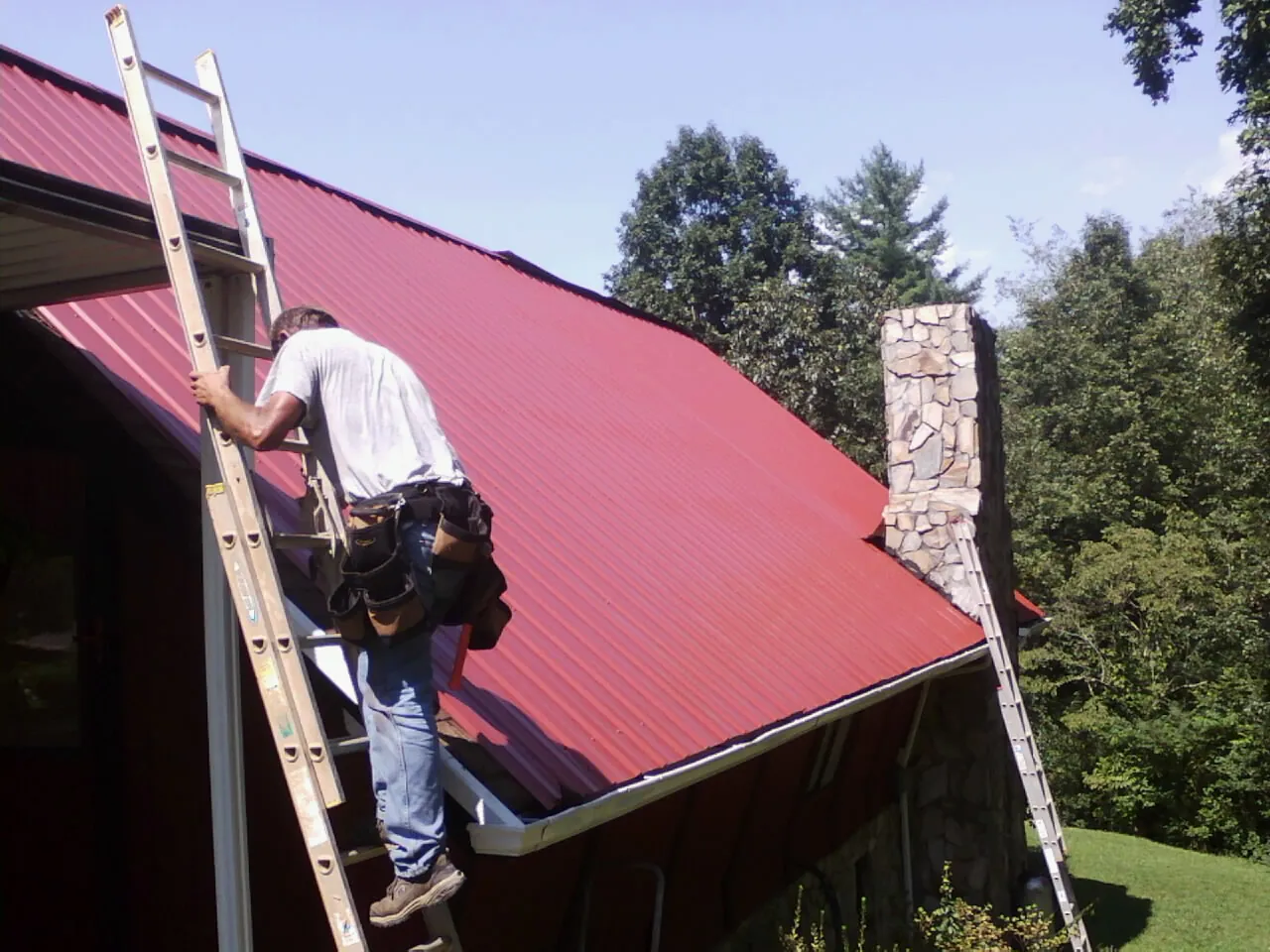 Roofer on a ladder beside a red metal roof and stone chimney — Soffit & Fascia Repair by Roofing Brothers in Blairs Mills